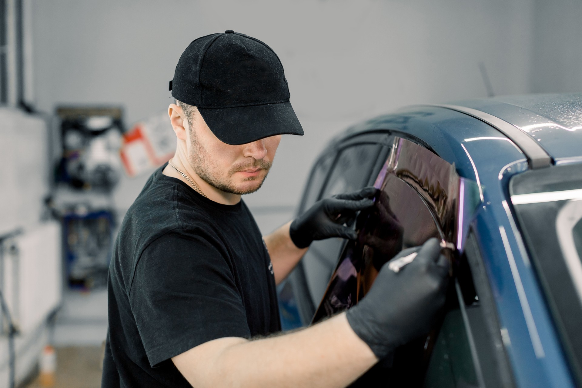 Closeup image of a handsome car mechanic worker, wearing black uniform, attaching tinting foil to car window in specialized service station
